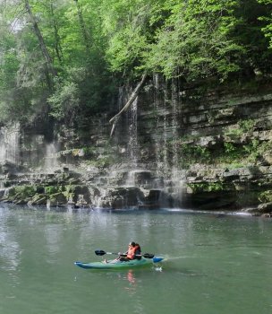A person kayaking on a calm river near a mossy, rocky cliff with cascading waterfalls. Surrounding lush green foliage creates a serene atmosphere.