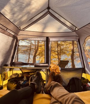 A person relaxes in a hammock near a campfire, surrounded by autumn trees. A lake reflects the warm sunset, creating a serene and peaceful atmosphere.
