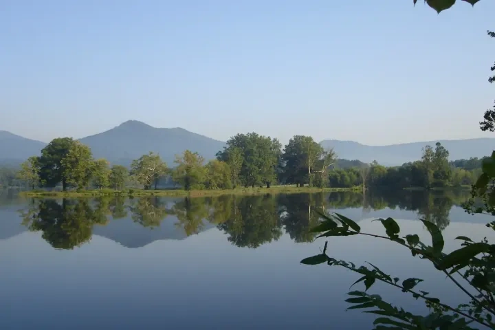 Calm lake reflecting trees and distant mountains under a clear blue sky.