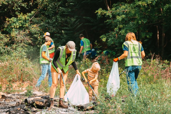 A group of volunteers, including a child, clean a wooded area. They're wearing green vests and using trash bags, conveying teamwork and environmental care.