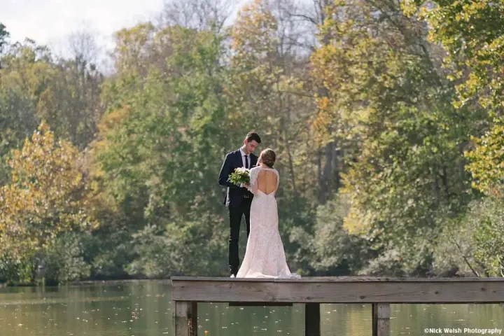 Bride and groom stand on a wooden dock, surrounded by trees and a calm lake.