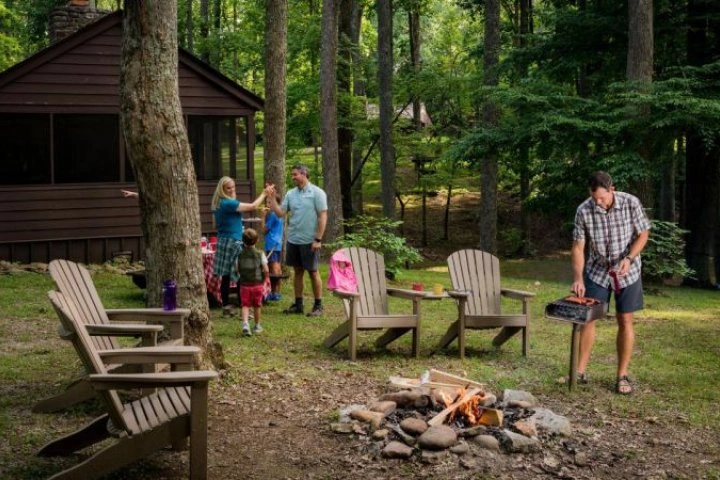 Family outside cabin in woods, man grilling by a campfire, chairs around.