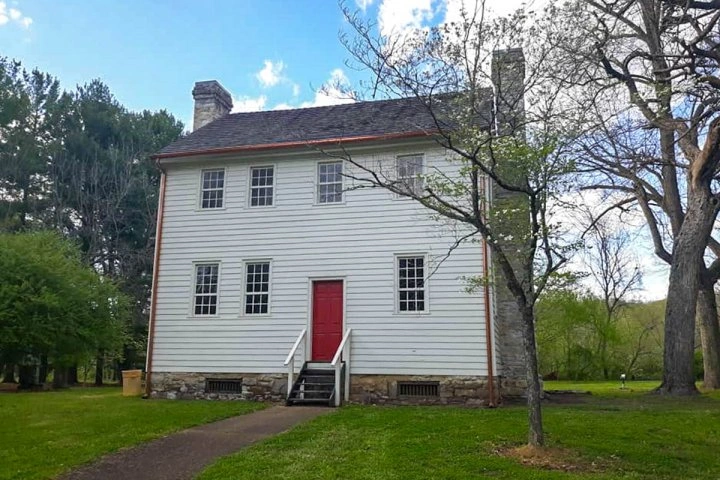 Historic two-story white wooden house with a red door, stone foundation, surrounded by lush green trees and grass under a partly cloudy sky.