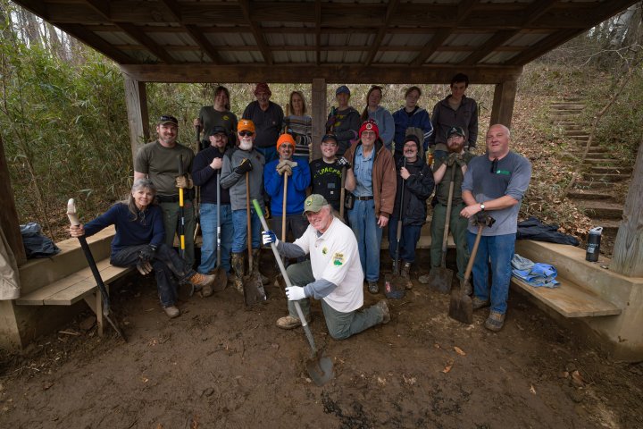 A group of 16 people, smiling and holding tools, poses under a wooden shelter in a wooded area. The mood is upbeat and collaborative.