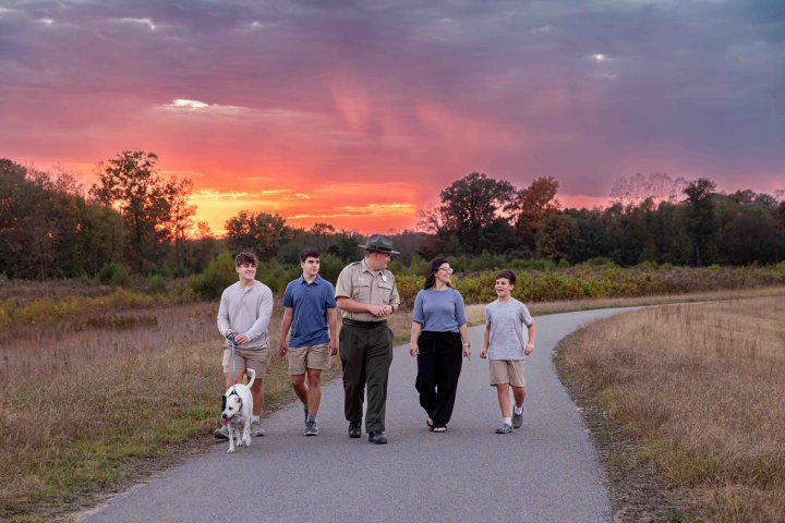 family with a dog and a ranger walking down a paved trail while the sun it setting
