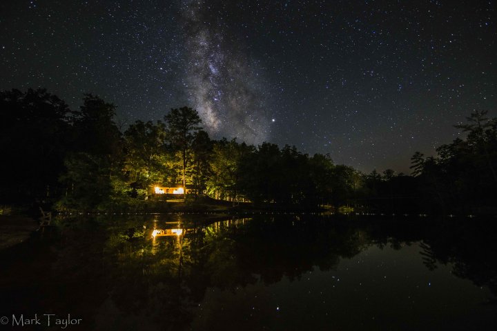 a campfire in the distance across a lake with the milky way in the night sky