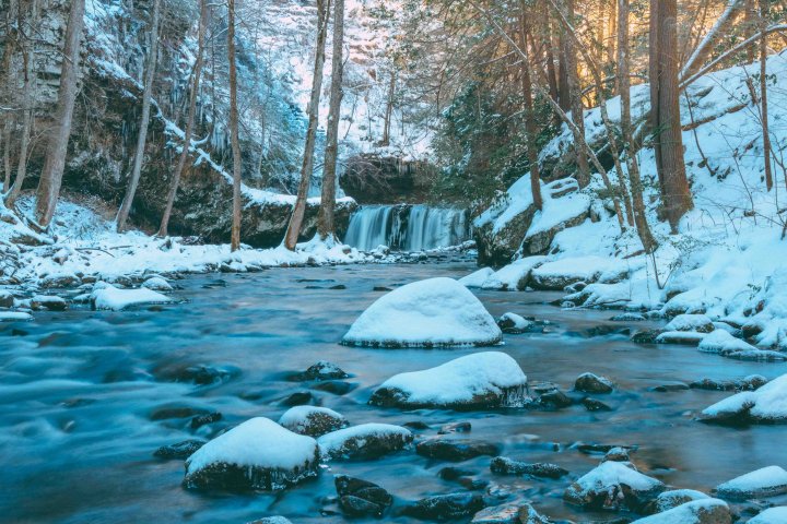 Snow-covered river with rocks and a small waterfall in a forest.