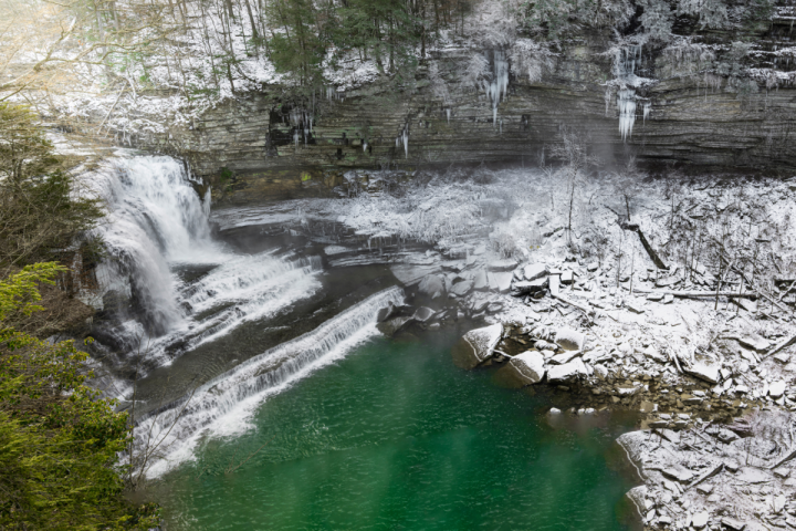 A large tiered waterfall spills into a blue pool surrounded by snow rocks and tall gorge walls