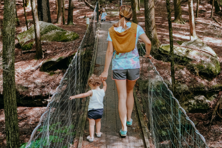 A mother and small child hold hands while crossing a suspension bridge in the woods.