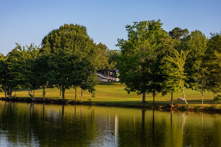 RV at a campsite among trees by a calm lakeshore with reflections in the water.