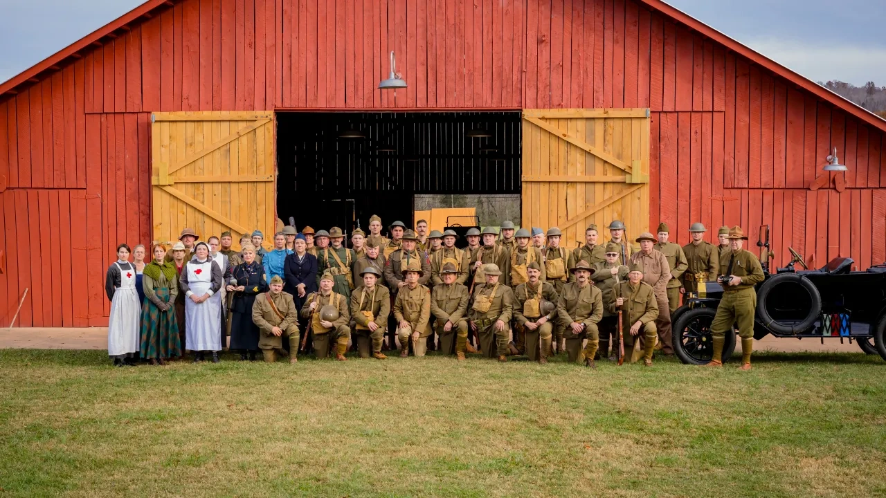 Group photo at a vintage reenactment with people in historical military uniforms and dresses in front of a large red barn, conveying nostalgia.