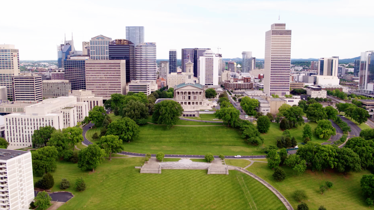 Aerial view of a cityscape featuring a prominent neoclassical building, surrounded by lush green lawns and trees. Tall skyscrapers in the background under a clear sky convey a lively urban atmosphere.