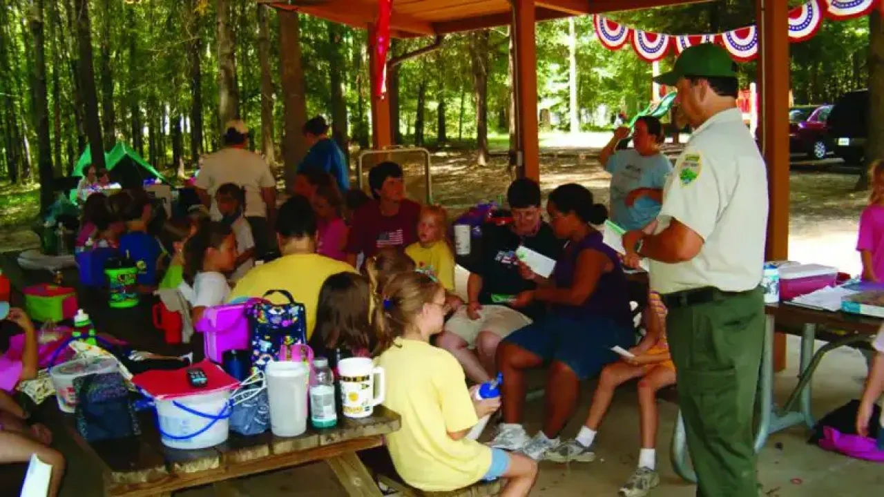 Families gather at a park pavilion for a summer picnic. A ranger in uniform talks to the group. Children and adults sit at tables, surrounded by trees.