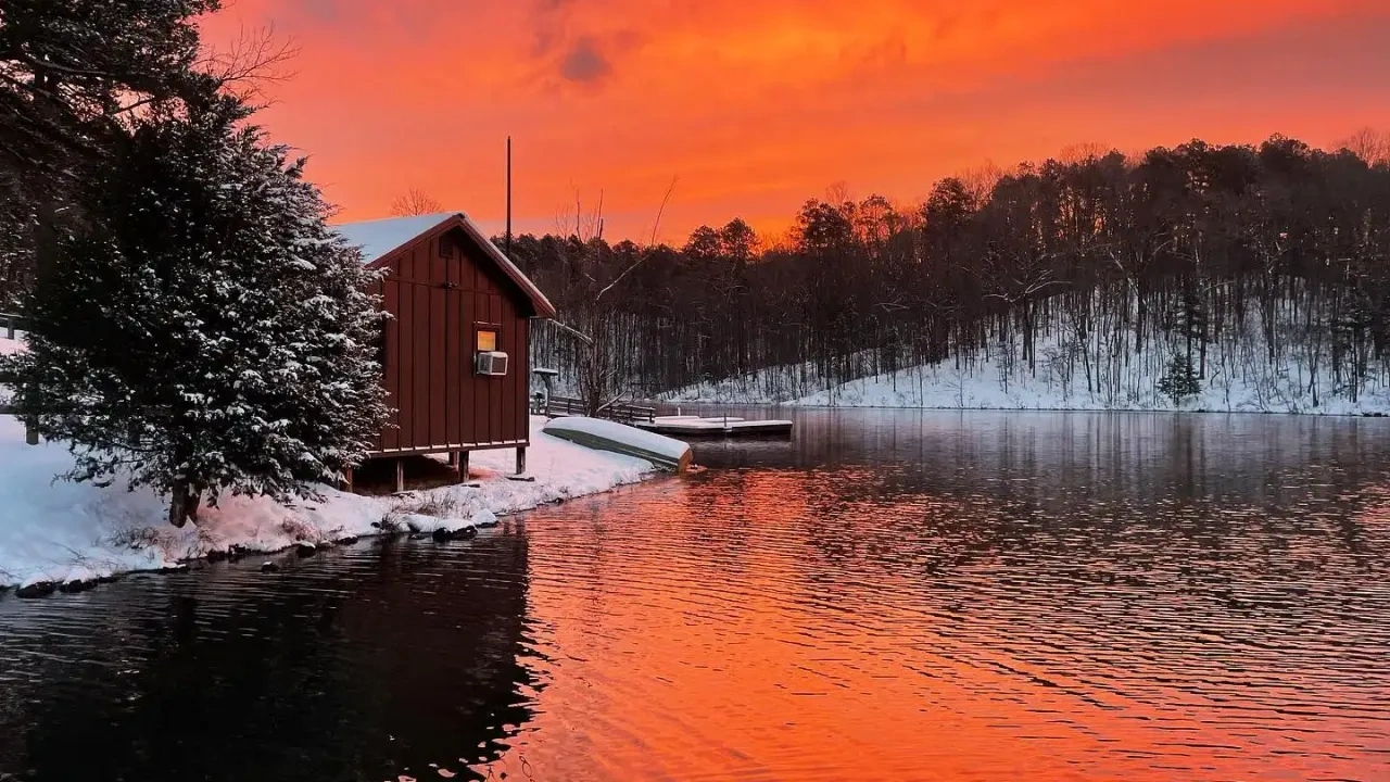 A snowy lakeside cabin with a vibrant, fiery orange sky reflecting in the calm water. Snow-covered trees line the horizon, creating a serene winter scene.