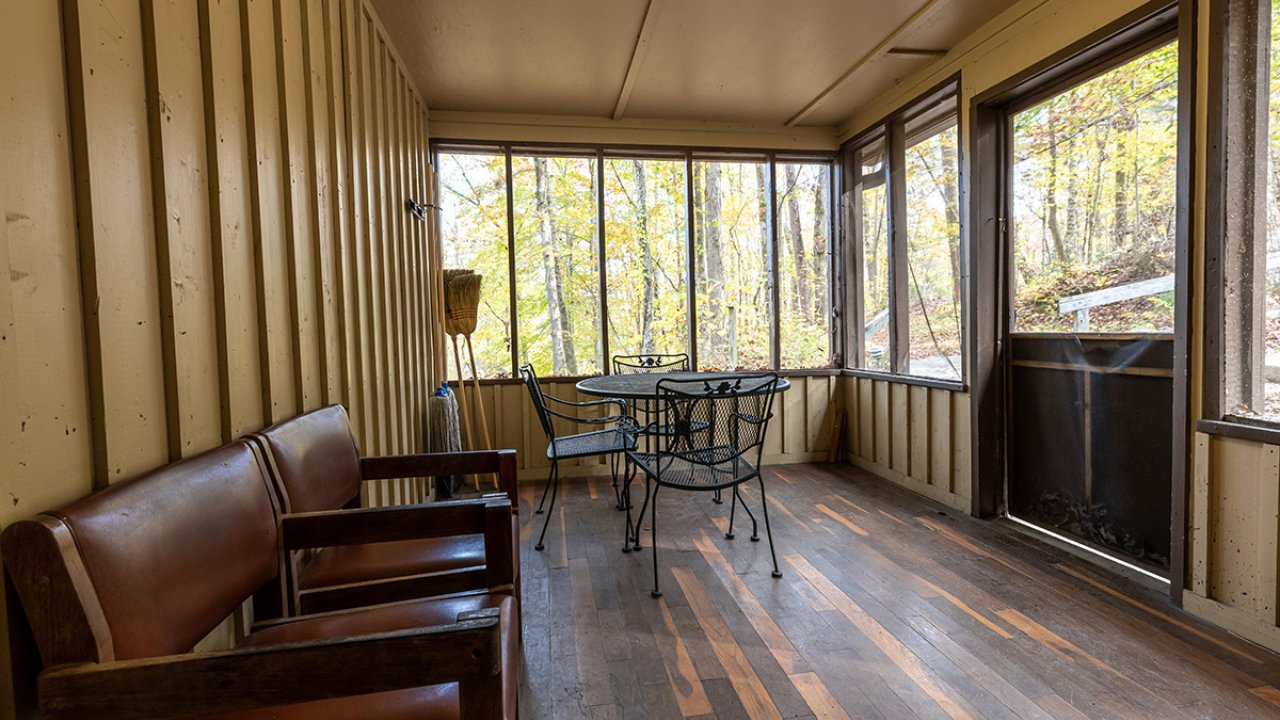 Breezeway inside a cabin on a sunny day with two chairs, a dinig room table and a screened in wall. 