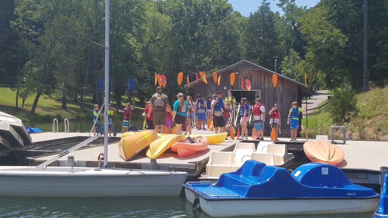 Group of people in life jackets hold paddles, standing on a dock by colorful kayaks and paddle boats. A wooden building and trees are in the background.