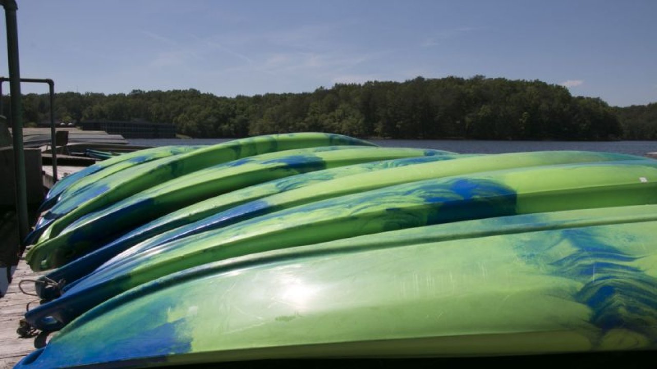 Alt text: "A row of green and blue kayaks stacked on a wooden dock beside a calm lake, with a backdrop of lush trees under a clear blue sky, conveying a serene and outdoor vibe."