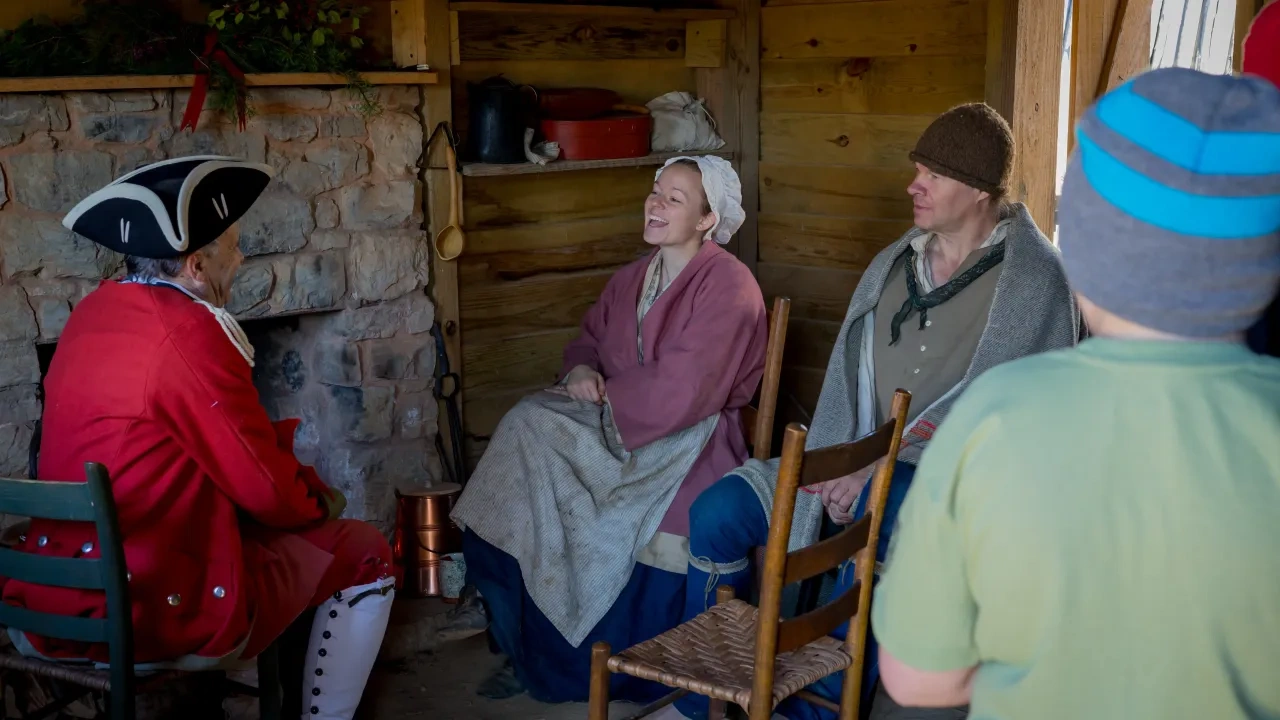 A group in historical attire sits in a rustic wooden cabin by a stone fireplace. They appear to be engaging in lively, cheerful conversation.