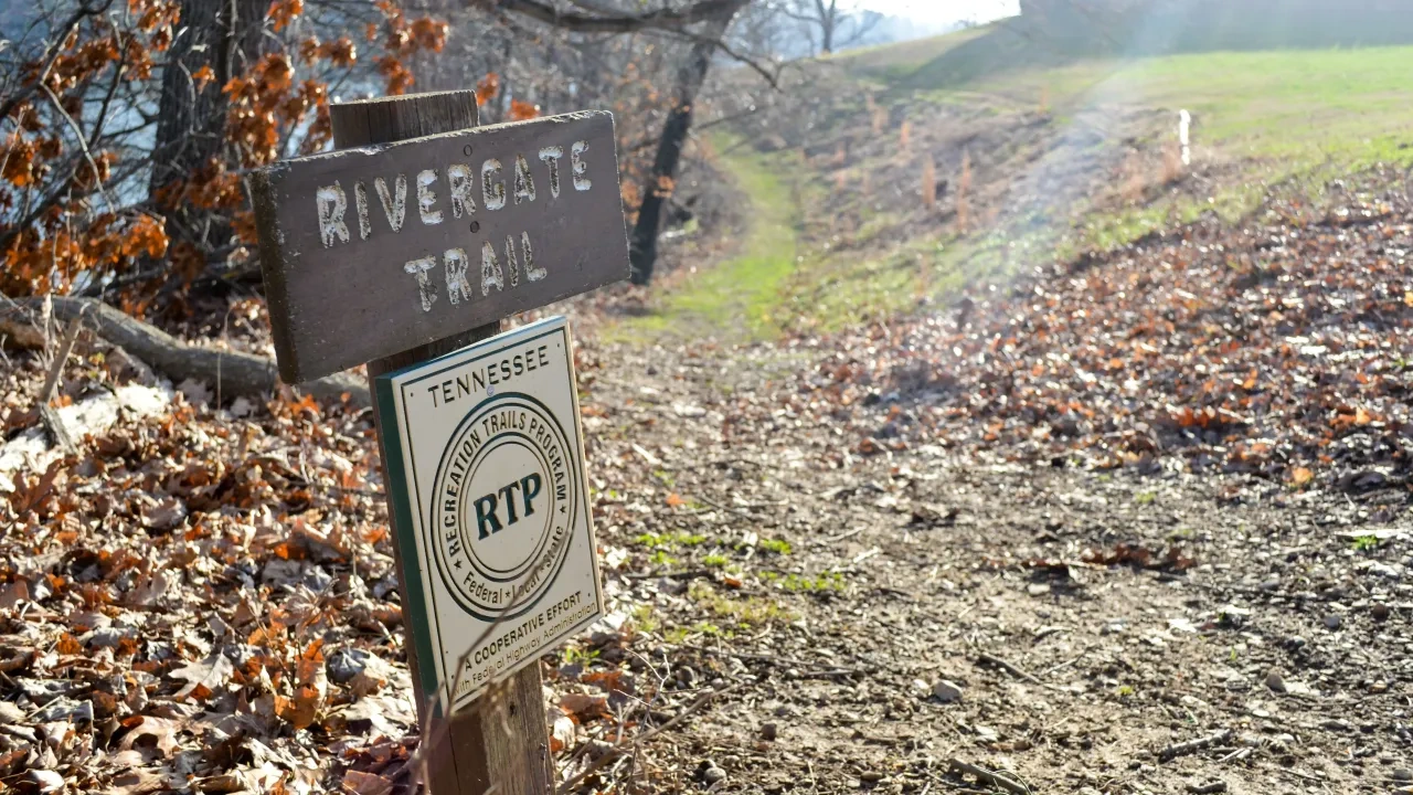 Trail sign in autumn leaves marks the Rivergate Trail in Tennessee. Sunlit path leads uphill, surrounded by bare trees and grassy patches.