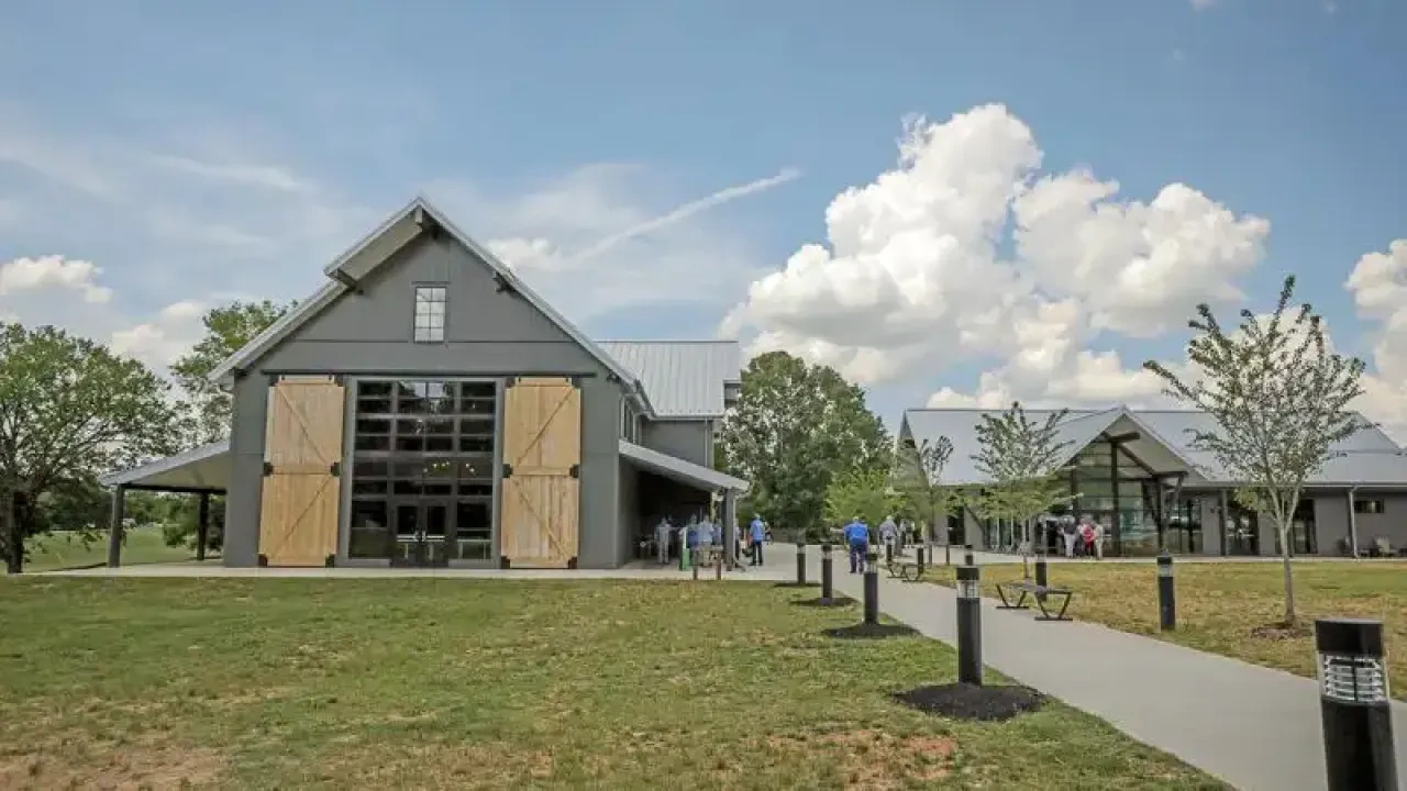 A modern barn-style building with large wooden doors set in a grassy landscape under a blue sky with fluffy clouds. People walk along a concrete path.