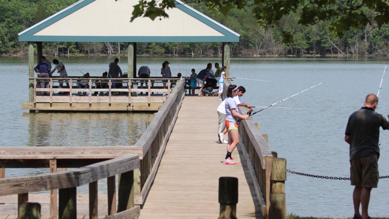 People are fishing from a wooden pier extending to a gazebo with a green roof is at the pier's end.