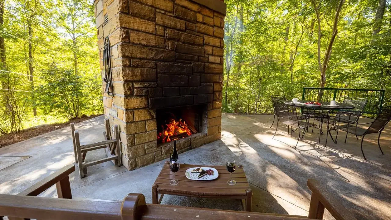 Outdoor patio with fireplace, wooden chairs, and dining table surrounded by lush trees.