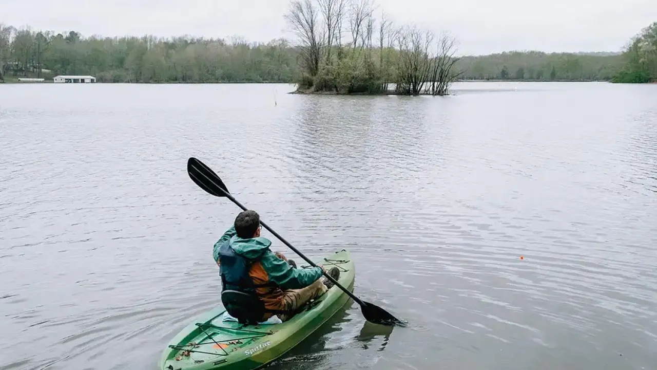 A person paddles a green kayak on a calm lake toward a small island with leafless trees, under an overcast sky.