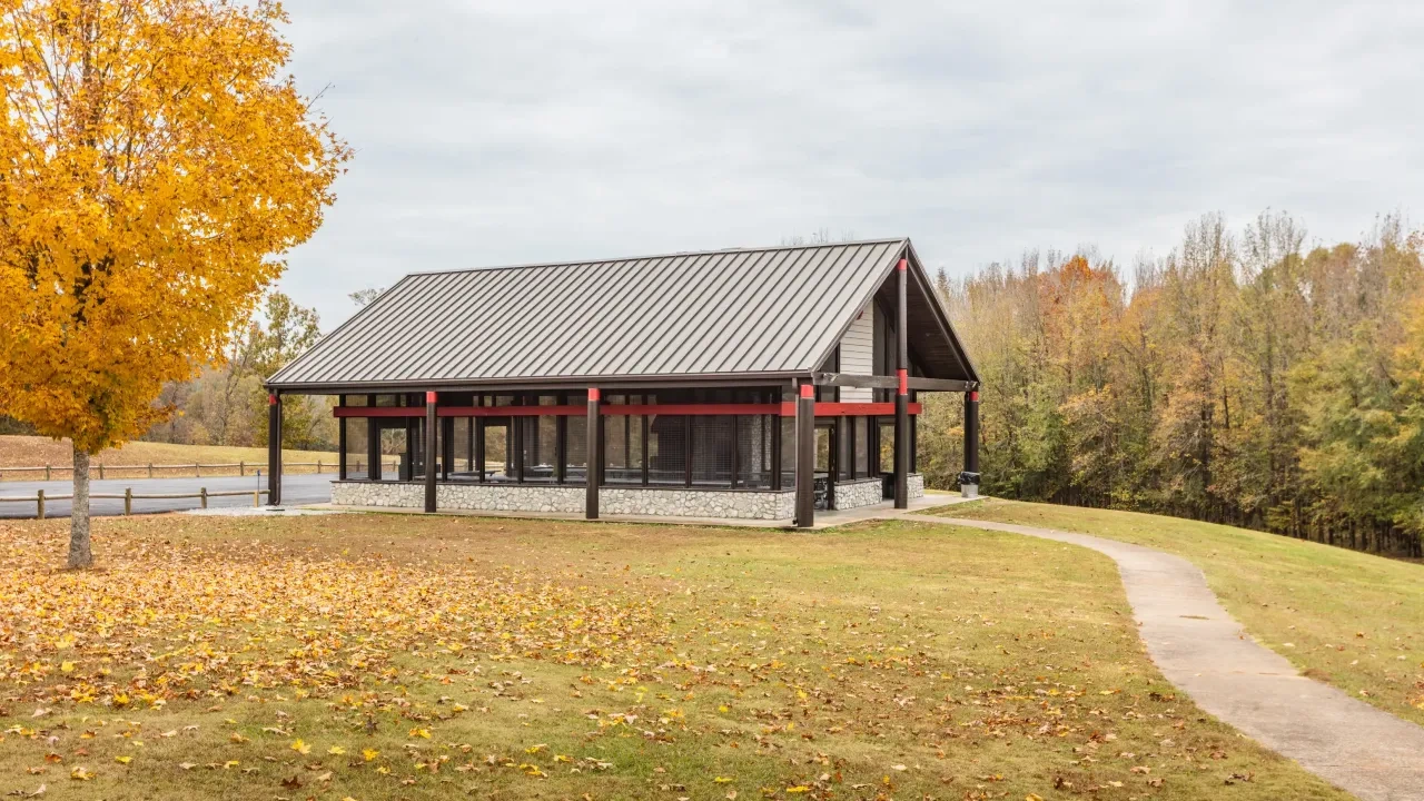 A modern pavilion building with a metal roof, stone base, and red trim stands on a grassy hill surrounded by autumn trees and a curving sidewalk.