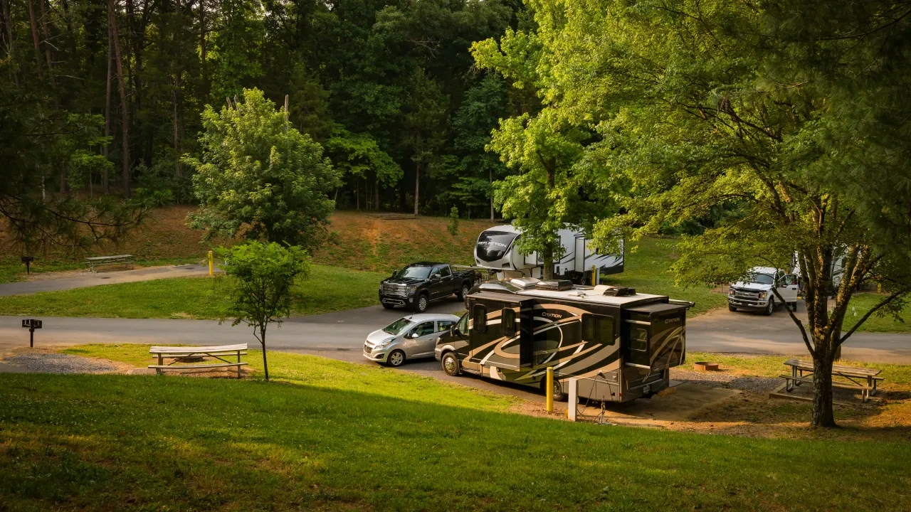 Campground scene with parked RVs and vehicles, surrounded by lush green trees.