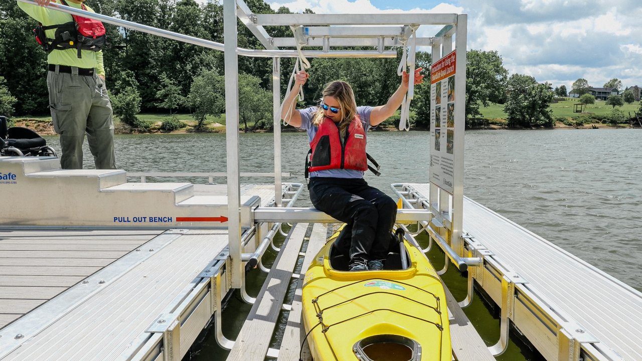 A woman in a life vest sits in a yellow kayak, using a launch mechanism on a dock by a lake. She appears focused, with green trees and cloudy skies in the background.