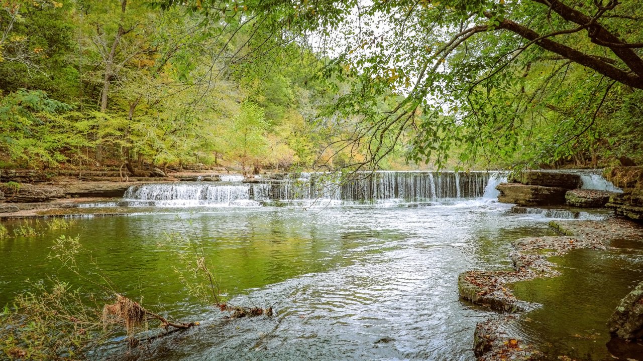 Tranquil scene of a small waterfall flowing into a clear river surrounded by lush, green trees. The calm water reflects the vibrant greenery above.