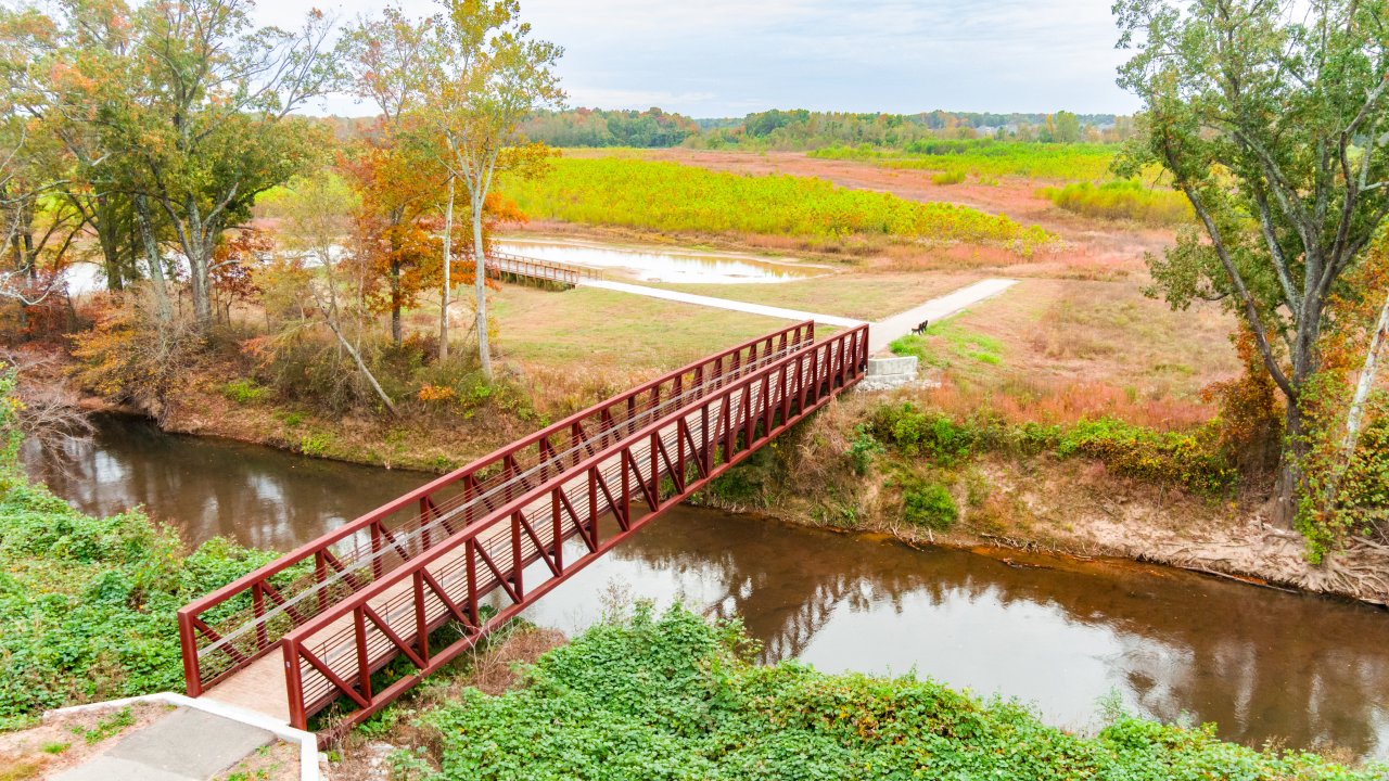 A red pedestrian bridge spans a calm river, surrounded by lush trees with autumn foliage and expansive, colorful fields under a cloudy sky.