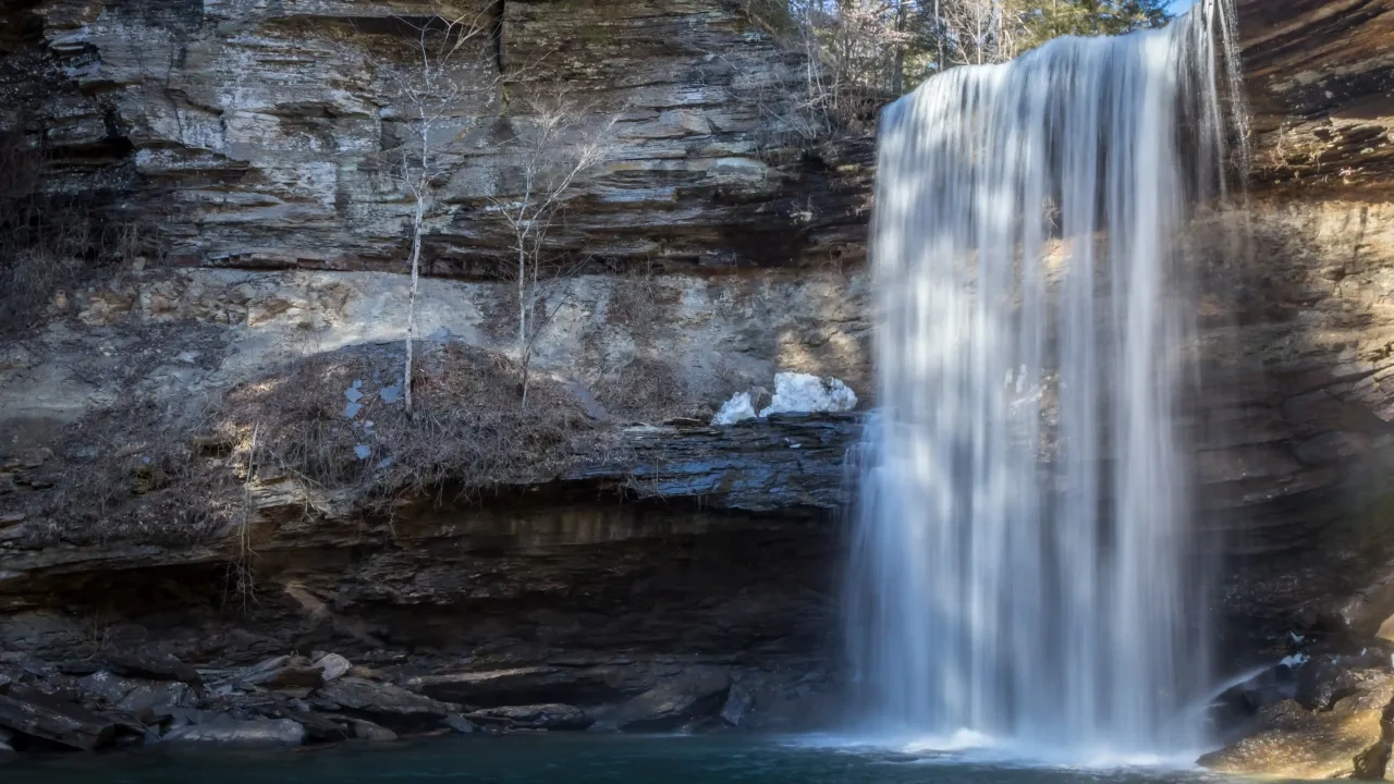 A serene waterfall cascades over a rocky cliff into a calm pool below. Sunlight illuminates the cliff, highlighting the texture of the rocks and creating a peaceful atmosphere.