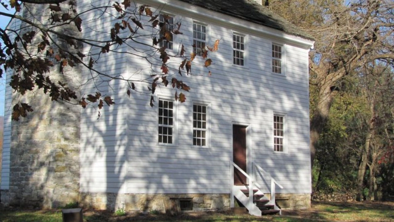 Historic white clapboard house with narrow windows, stone chimney, and small steps. Surrounded by autumn trees, casting dappled shadows on walls. Tranquil setting.