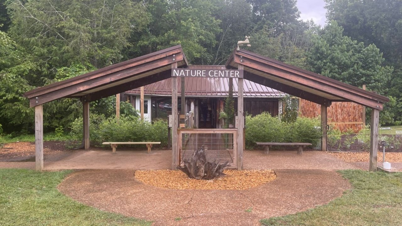 Wooden entrance with sloped roofs reading "Nature Center," surrounded by lush greenery and benches, creating a welcoming, serene outdoor atmosphere.