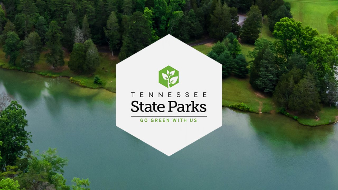 Aerial view of lush green forest beside a calm lake, overlaid with a hexagonal banner. Text reads: "Tennessee State Parks, Go Green With Us." Serene and inviting.