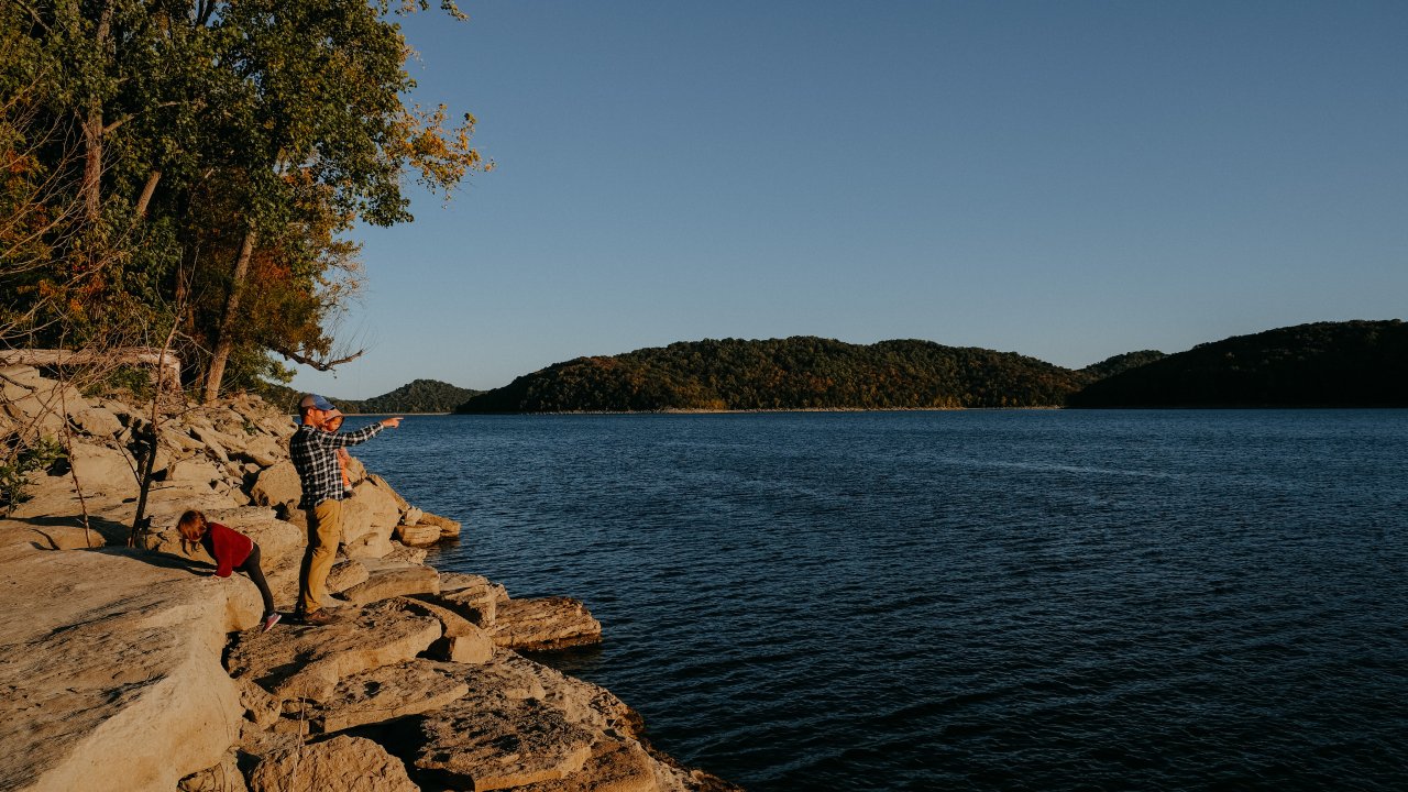 A person and a child stand on rocky lake shore, pointing across calm water towards distant forested hills. The scene is serene and bathed in warm sunlight.