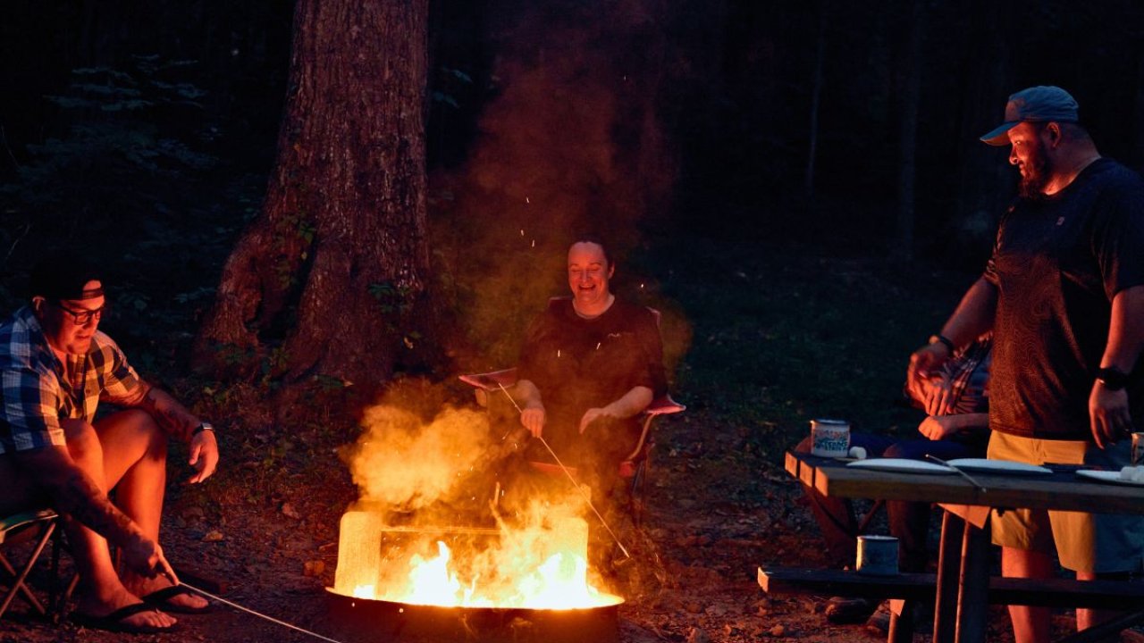 Three people enjoy a campfire at night in a forest, roasting food on sticks. The warm glow of the fire contrasts with the dark surroundings.