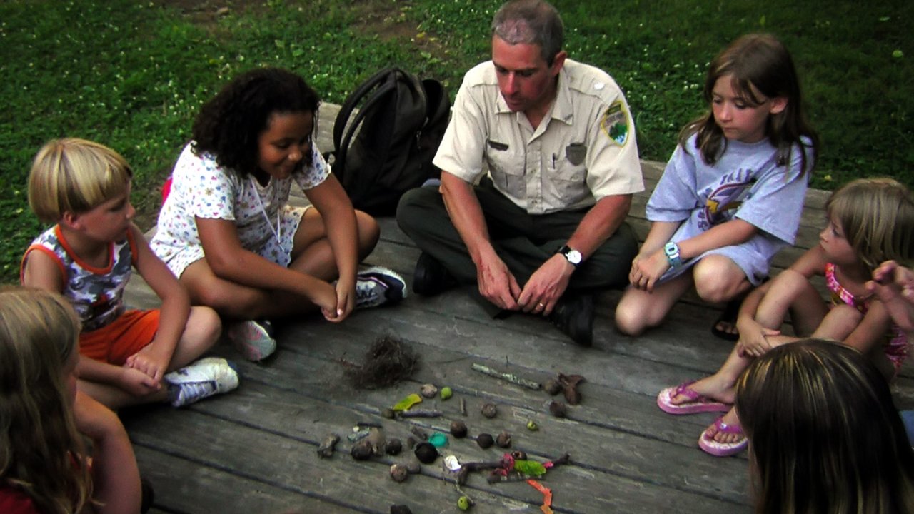 A park ranger sits on a wooden deck with a group of children, observing natural objects like rocks and sticks. The scene conveys learning and curiosity.