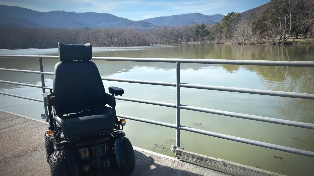 Motorized wheelchair by a lakeside pier, mountains in the background.