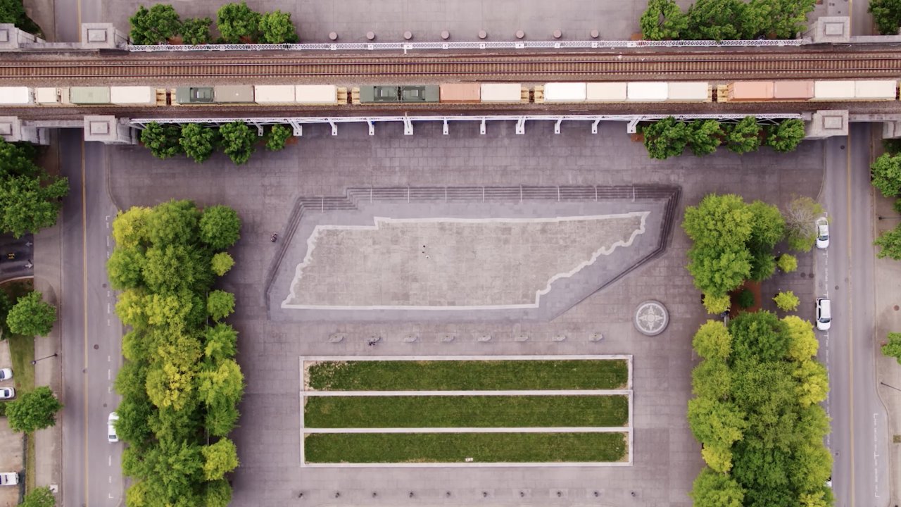 Aerial view of paved plaza featuring a large, outlined Tennessee shape. Train tracks run above, bordered by lush green trees lining the pathways.