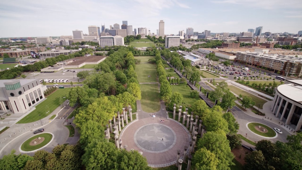 Aerial view of a city park with a circular plaza featuring a star pattern. Tree-lined paths extend towards a city skyline under a clear blue sky.