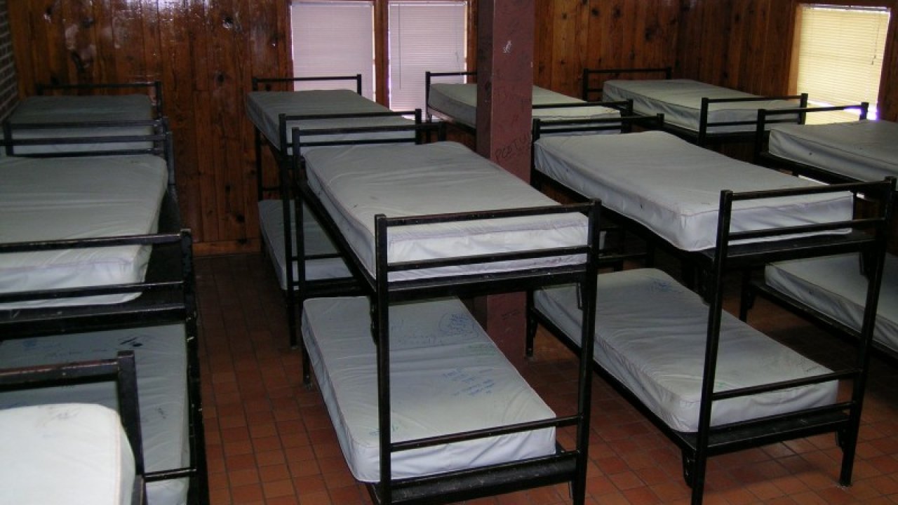 A dimly lit dormitory room with rows of simple metal bunk beds, wooden walls, and tiled floor, evoking a utilitarian and austere atmosphere.