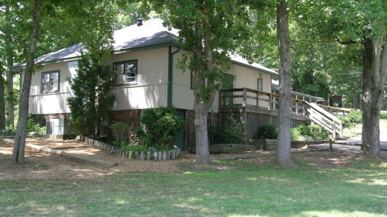 A cozy, single-story house with a light gray exterior and a sloped roof is nestled among tall trees, with a wooden ramp leading to the entrance. Sunlight filters through the leaves, casting dappled shadows on the grassy yard, creating a serene, inviting atmosphere.