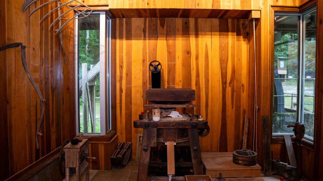 A rustic workshop with wooden walls features a central vintage printing press surrounded by various tools. Light streams through two side windows, creating a warm, historical ambiance.