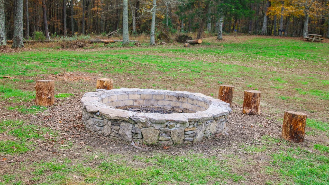 A stone fire pit with surrounding wooden log seats sits in an open grassy area, bordered by a forest of autumn-colored trees, evoking a cozy, rustic ambiance.
