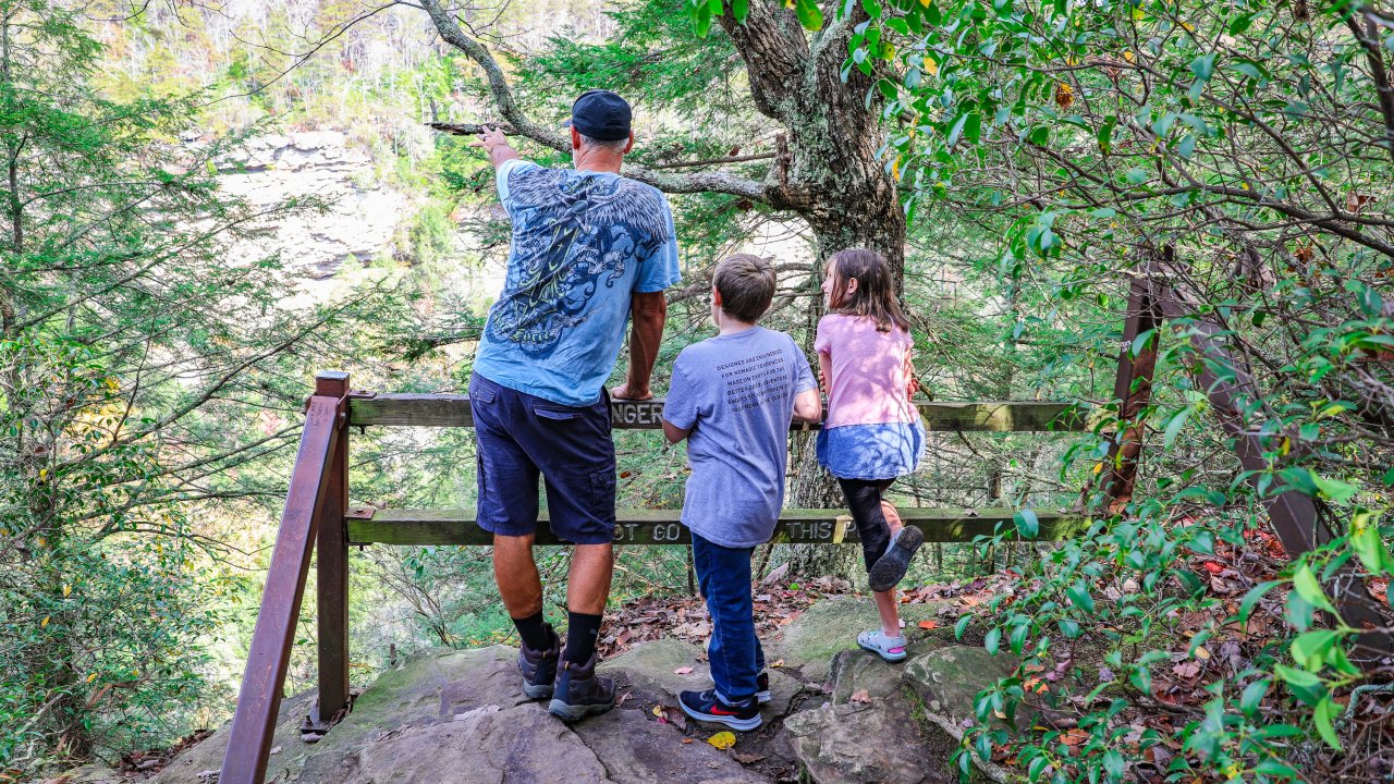 A man and two children in casual clothing stand on a rocky path in a lush forest, overlooking a scenic canyon with caution signs nearby.