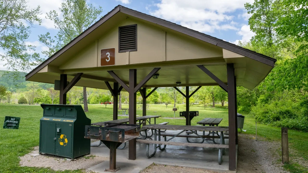 Park pavilion with picnic tables, grill, and recycling bins under a blue sky.
