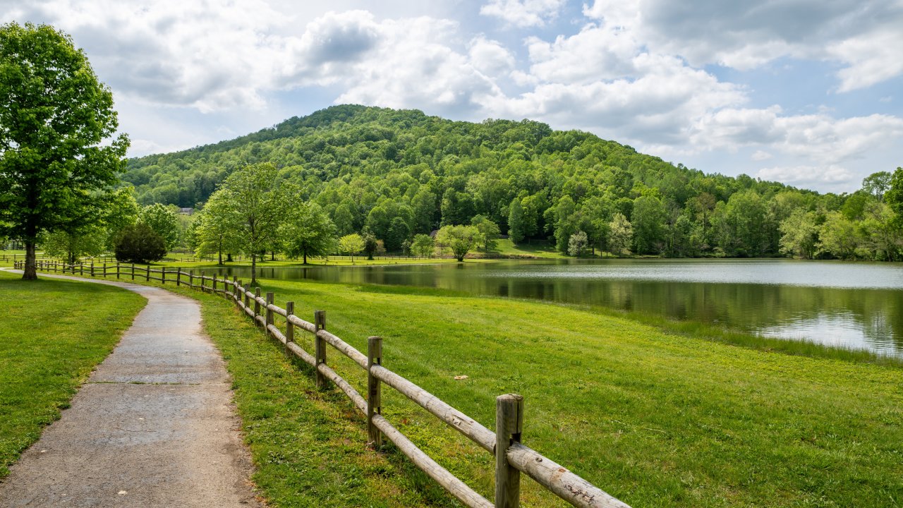 A winding path with a wooden fence leads to a lush green hill beside a tranquil lake under a partly cloudy sky, evoking a peaceful, serene atmosphere.