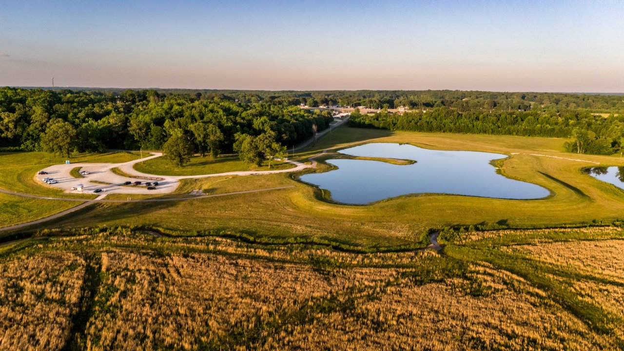 Aerial view of a tranquil park with a small lake, surrounded by green fields and dense forest. Cars are parked on a winding road. Sunny, peaceful setting.